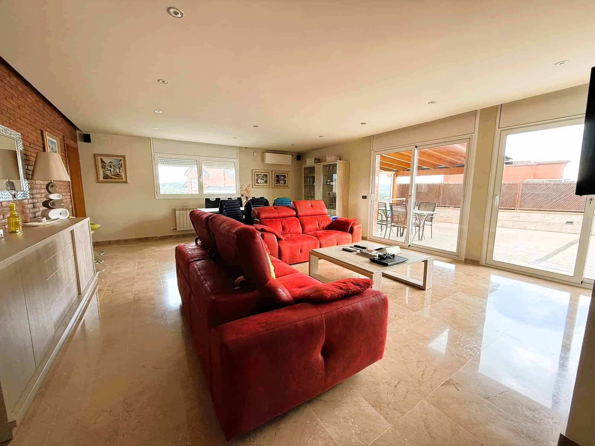 Amb interior ample i lluminós de casa amb mobiliari modern, sofàs vermells i grans obertures a la terrassa exterior, ideal per a serveis immobiliaris i assessorament legal en immobiliària.