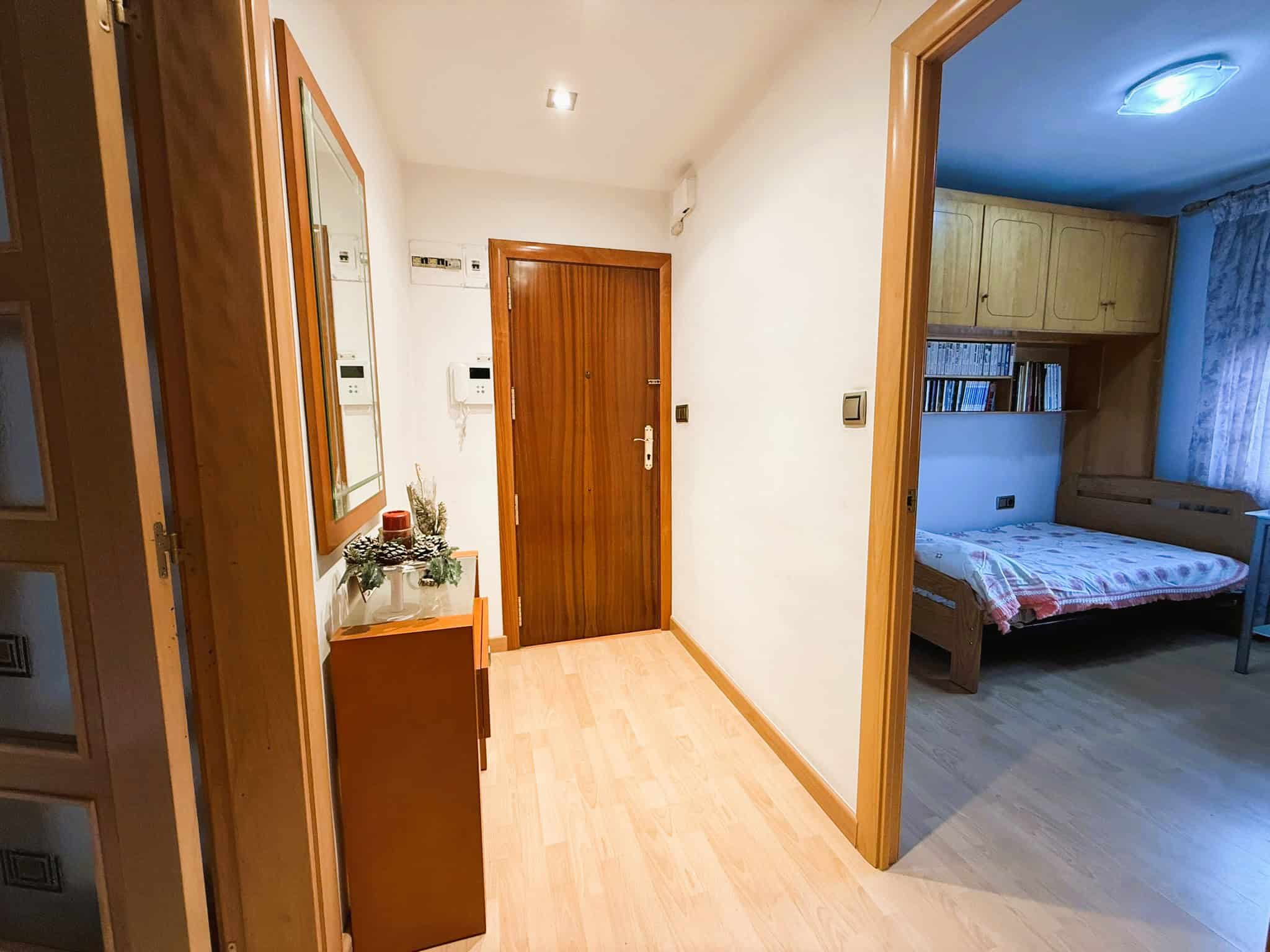 Mobilia d'interior d'un pis amb passadís blanc i portes de fusta, habitació amb llit de fusta i prestatge de llibres, decoració nadalenca a la consola.