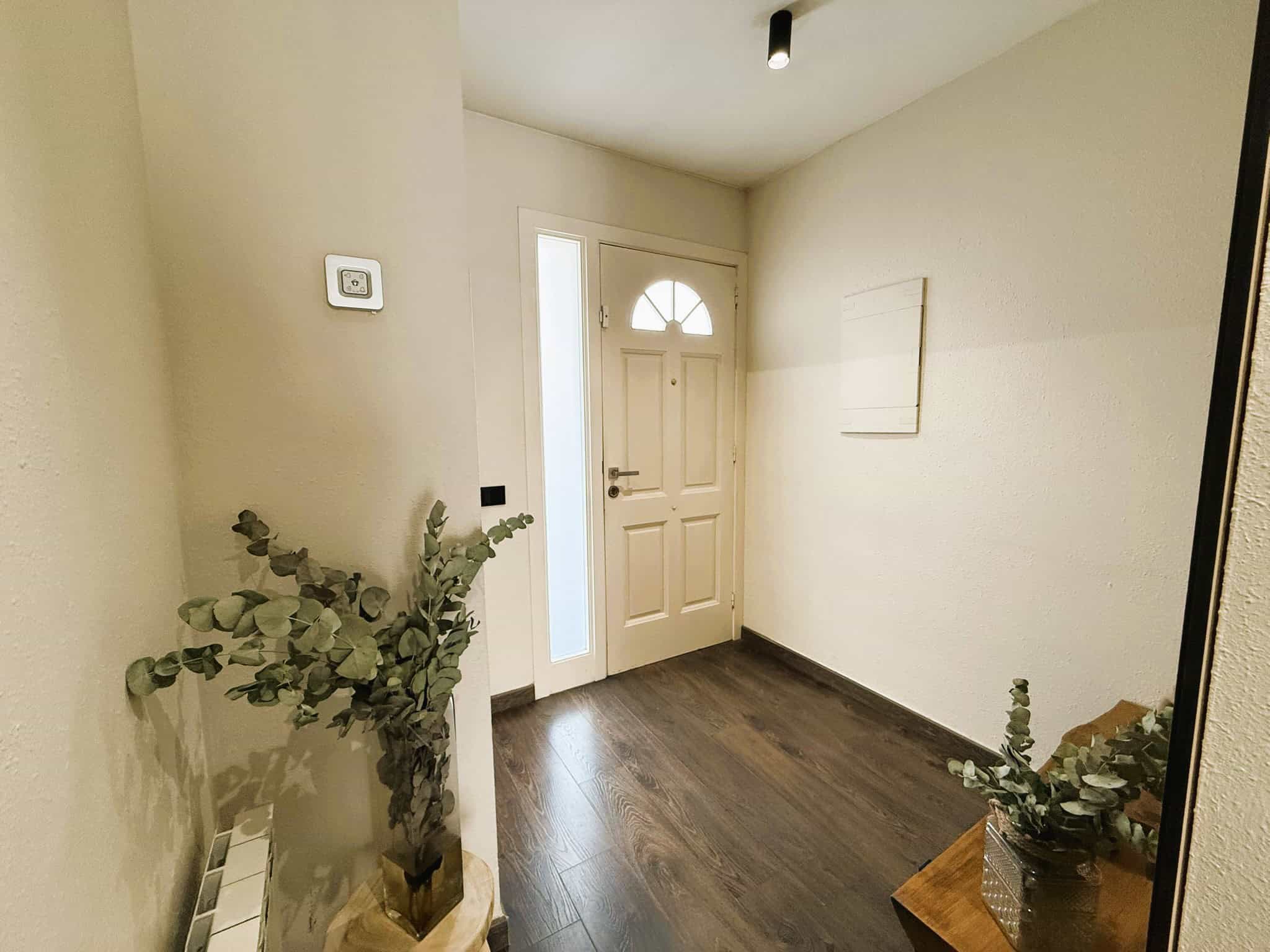 Imatge d'una entrada acollidora amb porta de fusta i finestra de vidre, decorada amb plantes verdes i un sòl de fusta fosca, ideal per a la venda o lloguer a Palau-solità i Plegamans.