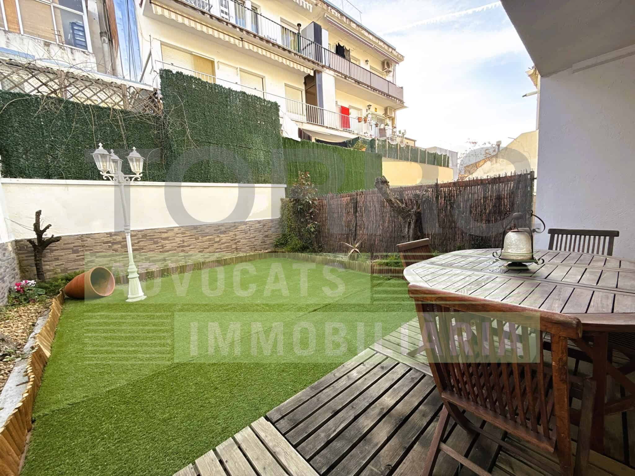Imatge d'una terrassa amb taula de fusta i cadires, jardí privat amb gespa i il·luminació exterior, envoltada de murs i vegetació, ideal per a activitats a l'aire lliure.